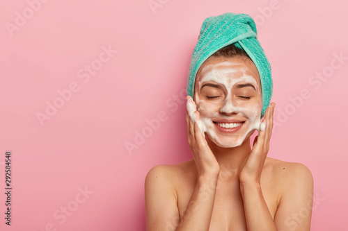 Pleased smiling woman washes face with cleansing gel, has soap on complexion, keeps eyes shut, wears wrapped towel on head, has naked body, poses against pink background, copy space on left.
