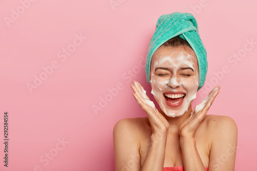 Happy joyous young girl spreads palms over face, washes face with soap, has fun in bathroom, pampers skin, wears wrapped towel on head, expresses positive emotions, poses against pink background
