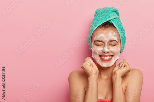 Joyful Caucasian woman clenches fists, smiles broadly, shows white teeth, washes face with soap, removes dirt, enjoys hygienic treatments at home, wrapped in soft towel, models topless indoor