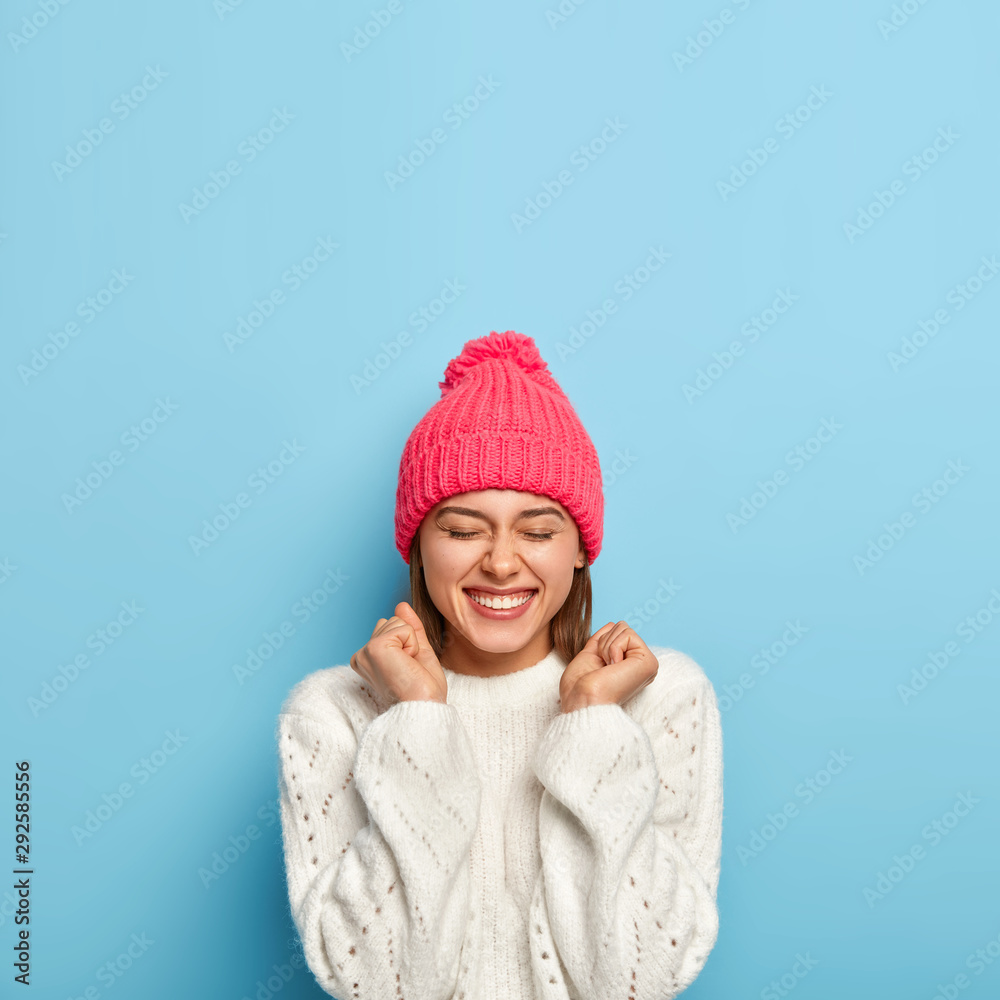 Vertical image of joyful young girl feels overjoyed, raises clenched fists, being in good mood, wears white sweater and pink hat, dressed in warm clothes during cold autumn day, isolated on blue wall