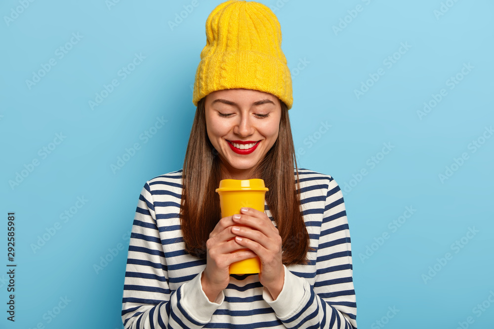 Horizontal shot of happy European woman wears stylish yellow hat and striped jumper, holds takeout coffee, has red painted lips, enjoys aromatic drink during outdoor walk with friends isolated on blue Horizontal shot of happy European woman wears stylish yellow hat and striped jumper, holds takeout coffee, has red painted lips, enjoys aromatic drink during outdoor walk with friends isolated on blue