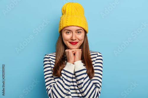 Image of tender good looking dark haired woman stands against blue background in yellow hat and striped sweater, wears red lipstick, makeup, looks staightly at camera. People, beauty, fashion