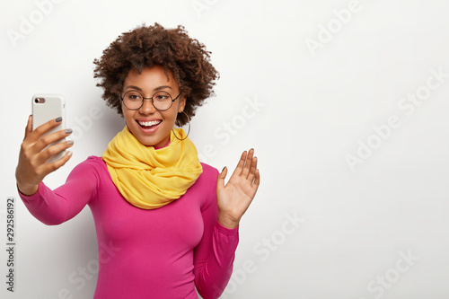 Smiling good looking Afro girl with crisp hair, waves palm in camera of smartphone, makes video call, wears transparent glasses and fashionable clothes, models against white background, copy space