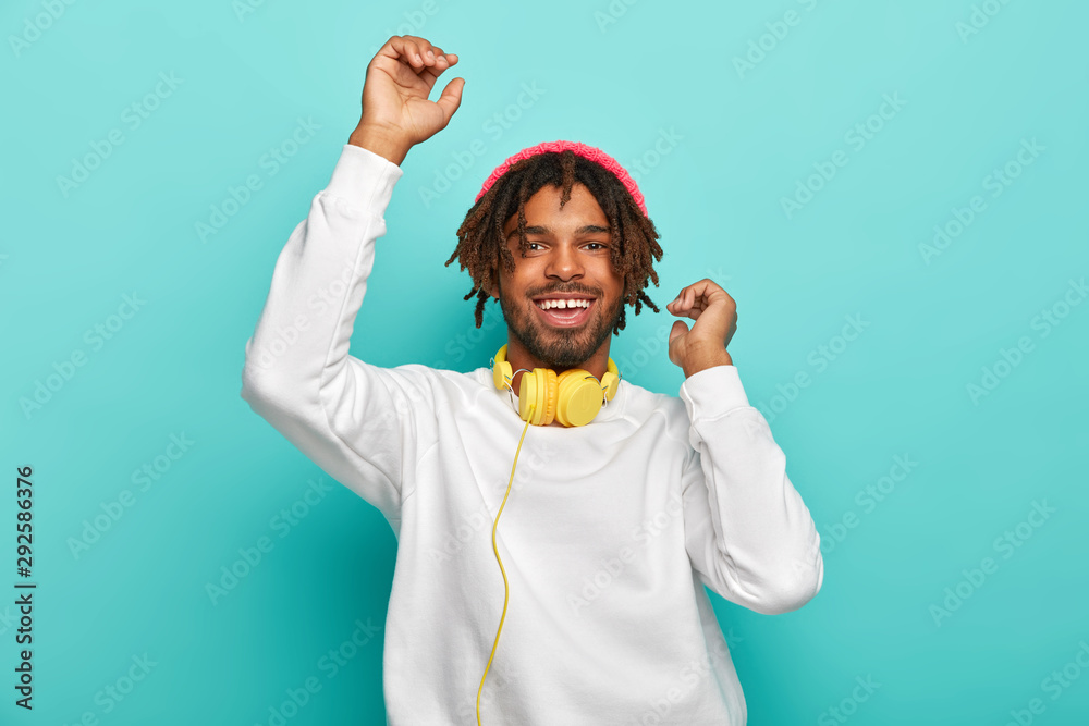 Upbeat carefree mixed race guy dances with hands up, listens awesome music track via headphones, has dreads, wears white comfortable jumper and stylish hat, isolated on blue. Time for fun and relax - obrazy, fototapety, plakaty 