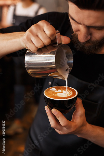 Young smiling male barista making cappuccino. Pouring milk for prepare cup of coffee. Latte art. Morning cup of coffee in café. Brewing coffee. Coffee shop concept. photo