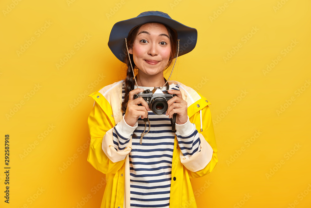 Smiling good looking Asian woman with long pigtail, wears hat, yellow raincoat, holds retro camera, takes pictures during her awesome trip, isolated over yellow background. Trekking, hobby concept