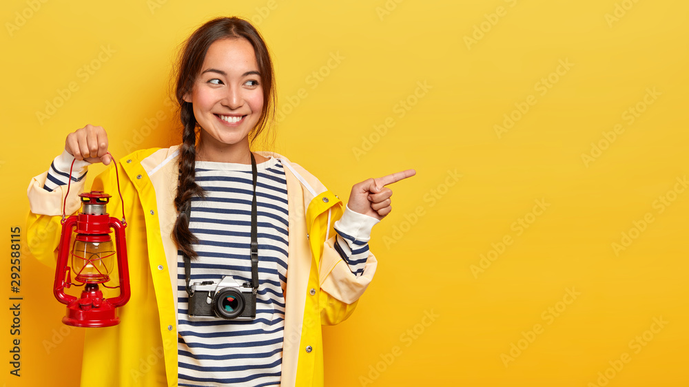 Obraz premium Positive Asian girl with tender smile, has dark hair, points aside on blank space, dressed in raincoat, holds gas lamp, enjoys adventurous trip, carries retro camera with her, smiles pleasantly