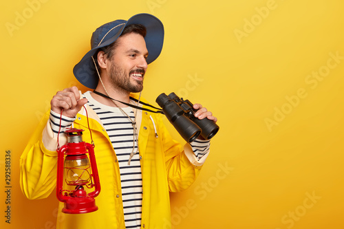 Adventure time and summer travel concept. Happy male traveler has outdoor activities, explores world, uses binoculars and torch dressed in waterproof raincoat leads active lifestyle isolated on yellow photo