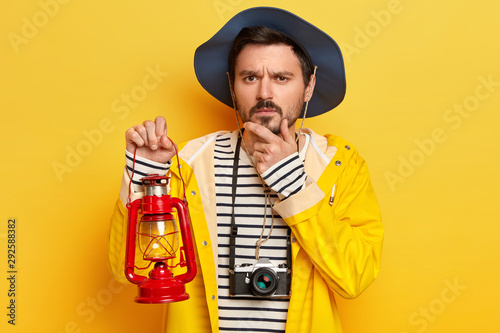Studio shot of serious man holds chin, thinks about something, carries kerosene lamp, has retro camera hanging around neck, enjoys travel in mountains or forest, dressed in raincoat isolated on yellow photo