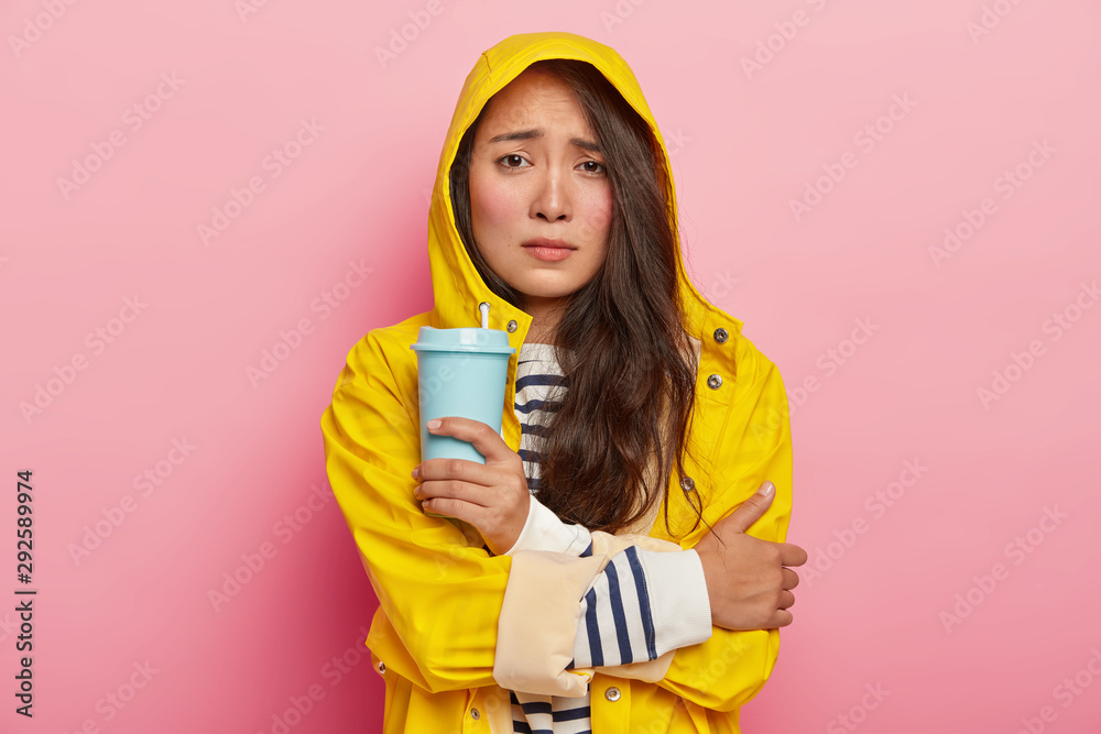 Photo of dissatisfied Asian woman smirks face, keeps arms crossed, trembles from being cold after walking in rain, wears yellow raincoat, holds takeaway coffee, warms herself with hot beverage Photo of dissatisfied Asian woman smirks face, keeps arms crossed, trembles from being cold after walking in rain, wears yellow raincoat, holds takeaway coffee, warms herself with hot beverage