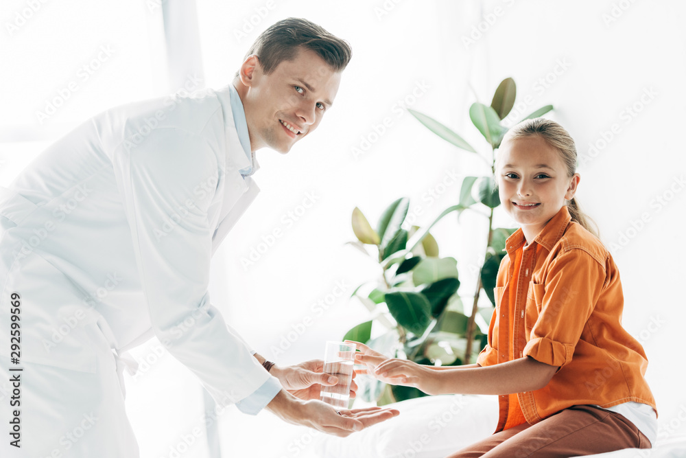 smiling pediatrist in white coat giving glass of water to child in clinic smiling pediatrist in white coat giving glass of water to child in clinic