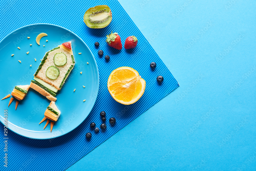 Fototapeta top view of plates with fancy rocket made of food on blue background with fruits