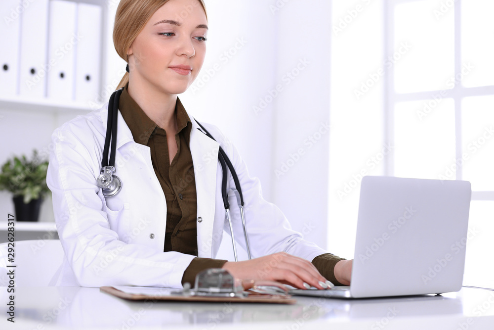 Young woman doctor at work in hospital looking at laptop pc monitor. Physician controls medication history records and exam results. Medicine and healthcare concept Young woman doctor at work in hospital looking at laptop pc monitor. Physician controls medication history records and exam results. Medicine and healthcare concept