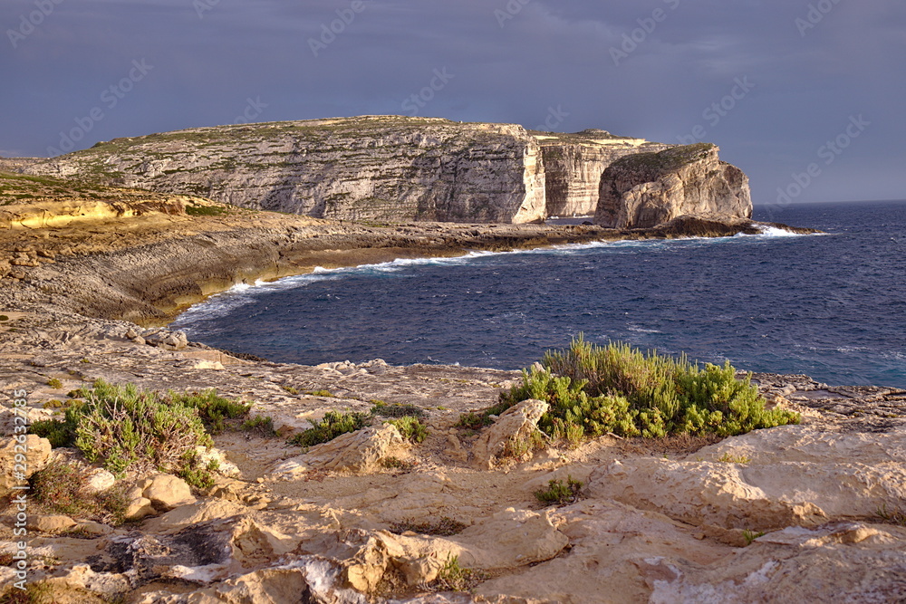 Beautiful round bay, green plants and fungus rock cliffs at sunset in gozo. Orange sunset light with grey storm clouds.