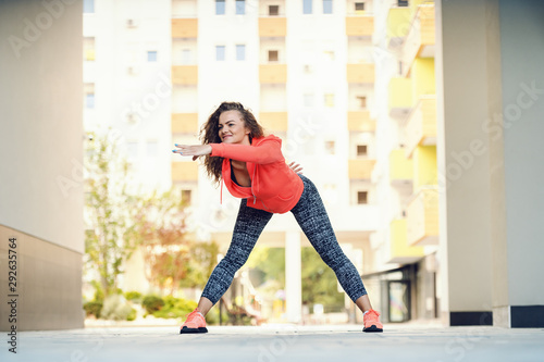 Focused attractive caucasian woman in sportswear and with curly hair bending with arm stretched and doing warm up exercises while standing wide-legged in passage on sunny day. photo