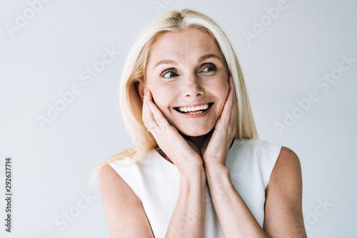 happy blonde mature woman with hands near face looking away isolated on grey