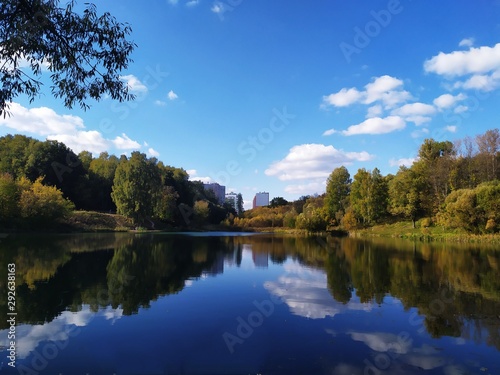Picturesque autumn landscape on the outskirts of the city. Multi-storey houses in the distance. Park with colorful trees and a lake. Reflection in the water. Rest and relaxation.