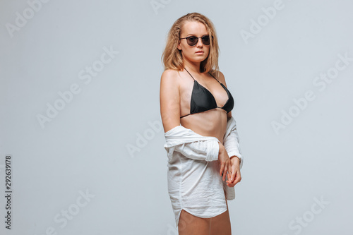Girl in a black swimsuit and sunglasses and a white shirt on a light gray background © Smeilov