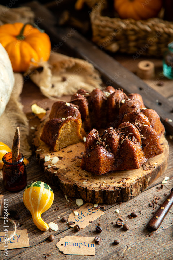 Fototapeta premium tasty homemade baked pumpkin bundt cake with chocolate on top stands on wooden board on rustic table with assorted colorful small pumpkins and autumn leaves in straw basket