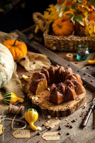 tasty homemade baked pumpkin bundt cake with chocolate on top stands on wooden board on rustic table with assorted colorful small pumpkins and autumn leaves in straw basket