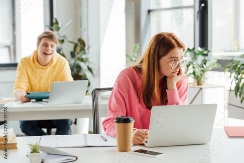 pensive girl in glasses using laptop while classmate laughing at her