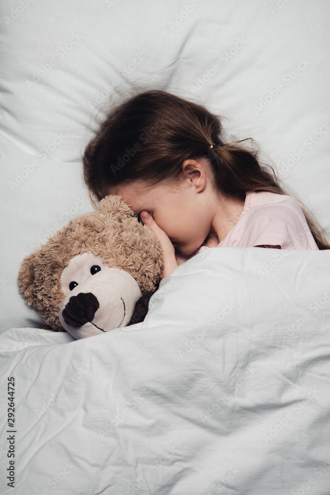 adorable child covering eyes with hand while lying in bed with teddy bear adorable child covering eyes with hand while lying in bed with teddy bear
