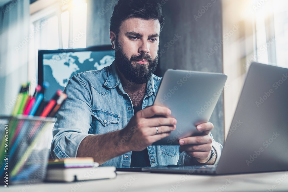 Professional at office desk using tablet in his work.Dark-haired man sitting at window in front of laptop with tablet in his hands