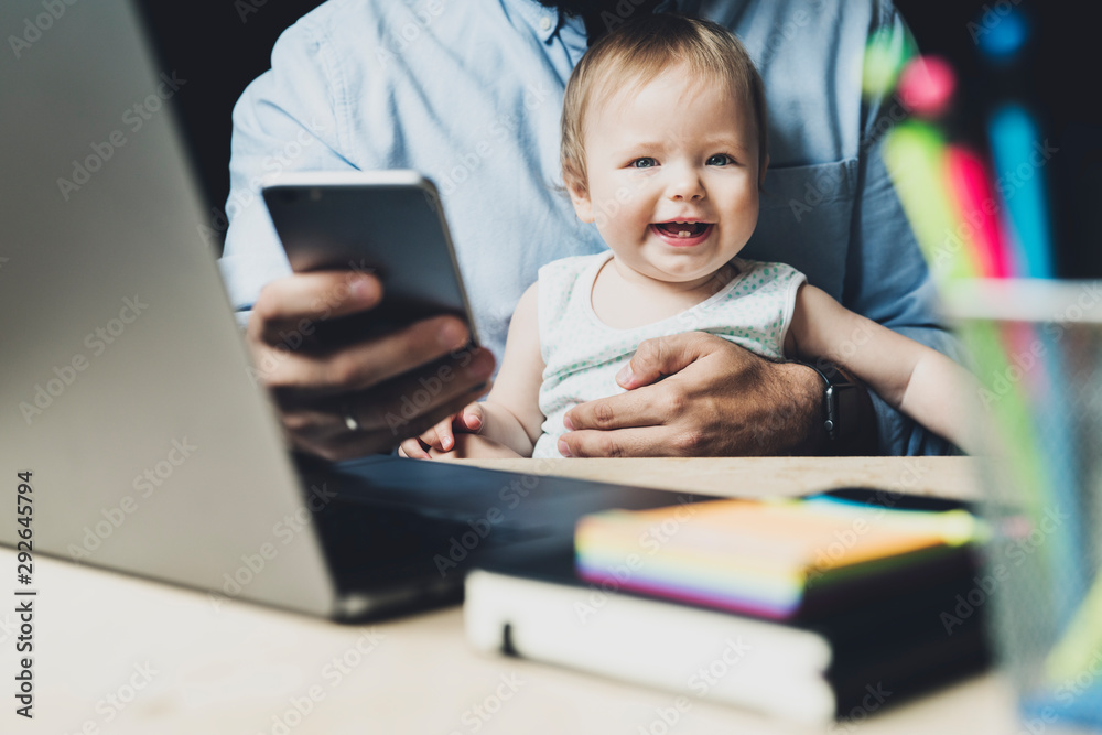 Little baby sitting on father's knees at his workplace. Man holding child in front of laptop and working remotely. Homebased job for young parents. Distant education for mothers and dads with infants. Little baby sitting on father's knees at his workplace. Man holding child in front of laptop and working remotely. Homebased job for young parents. Distant education for mothers and dads with infants.