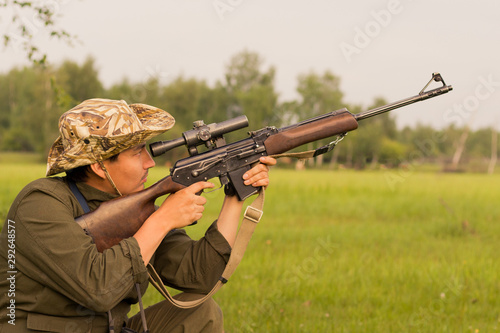 A male hunter with a gun while sitting takes aim at a forest. The concept of a successful hunt, an experienced hunter. Hunting the summer season. The hunter has a rifle and a hunting uniform. Image.