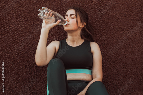 A young girl goes in for sports on the street and drinks water while sitting near the wall.