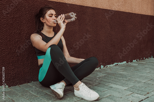 A young girl goes in for sports on the street and drinks water while sitting near the wall.