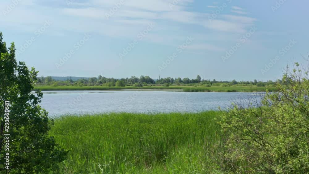 A drone flies from the shore of the lake past the bushes towards the village, crossing the lake. A strong wind sways grass and creates waves on the surface of the water. A drone flies from the shore of the lake past the bushes towards the village, crossing the lake. A strong wind sways grass and creates waves on the surface of the water.