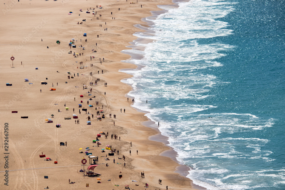 Drone view of people on the yellow sand beach facing the Beautiful crushing waves of Atlantic ocean in Nazare, Portugal