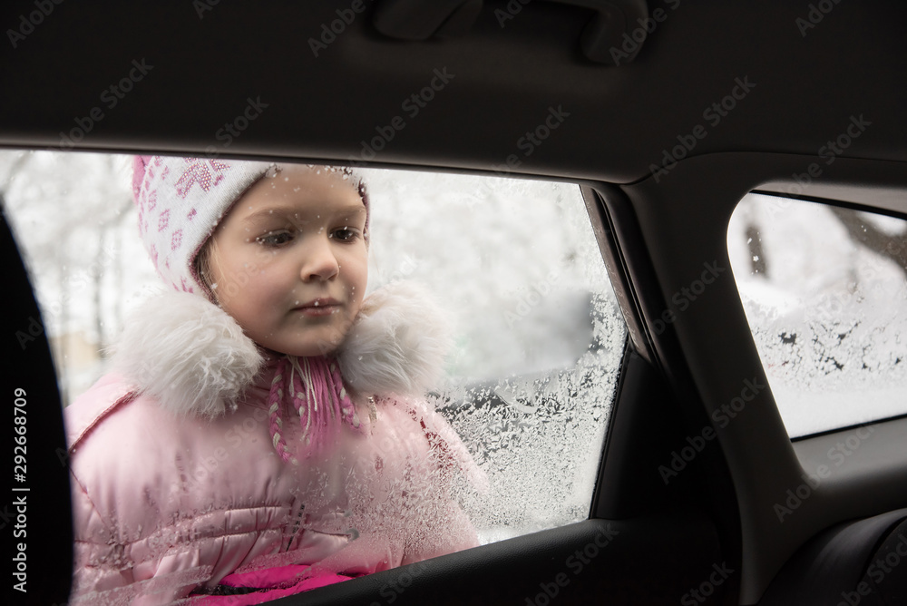 Little beautiful girl dressed in a warm pink jacket and hat, in winter, looks at snowflakes on a car window
