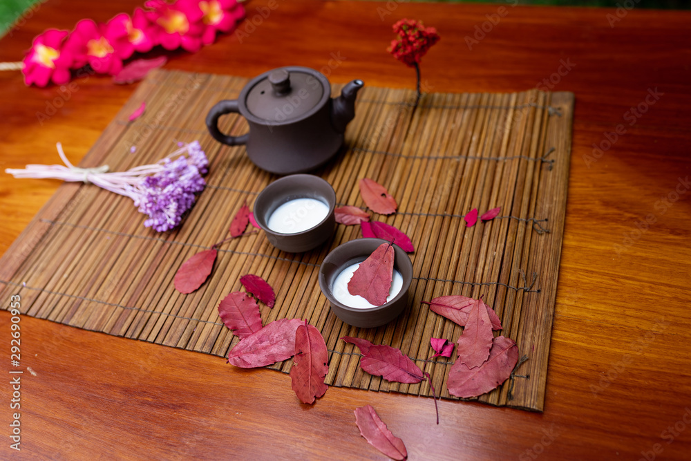 A small clay teapot with two cups for drinks standing on a mat among red sheets with a wooden sprig of lavender and red magnolia flowers