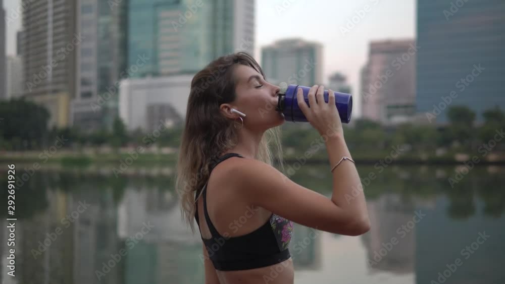 woman with airpods having drink from fitness blue cup after exercise in city park against high buildings woman with airpods having drink from fitness blue cup after exercise in city park against high buildings