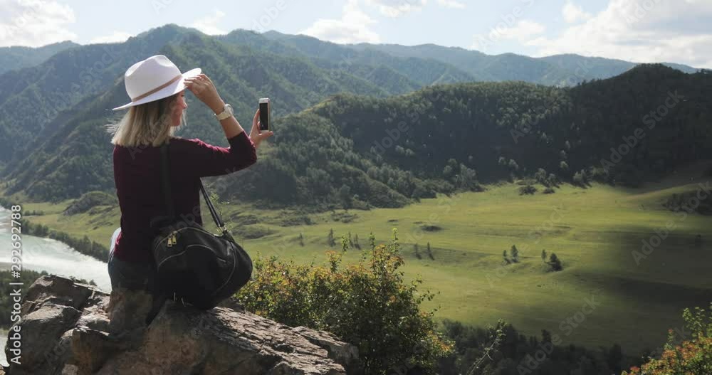 Making video and photo for blog using smartphone, modern freelance work. Woman traveler blogger shooting content on phone sitting on top of mountain in amazing valley. Eco tourism and travelling.