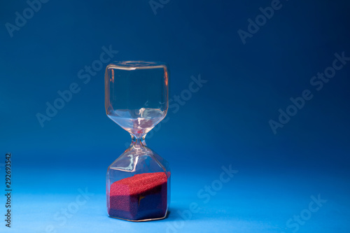 Fotografi Transparent hourglass with red grains of sand located on a blue background with free space for text