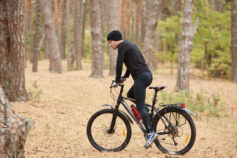 Side view of young European male riding bike in forest on sunny day, sporty man wearing black sportwear and cap onbicycle in wood, doing sport in open air. Sport, fitnes and healthy lifestyle concept.