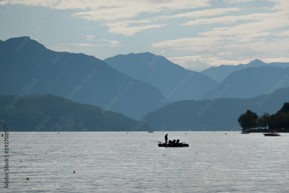 View of Lake Annecy. France. In the background are the Alps, hidden behind a blue haze. Silhouettes of boats are visible on the lake. Scenery. Background. View of Lake Annecy. France. In the background are the Alps, hidden behind a blue haze. Silhouettes of boats are visible on the lake. Scenery. Background.