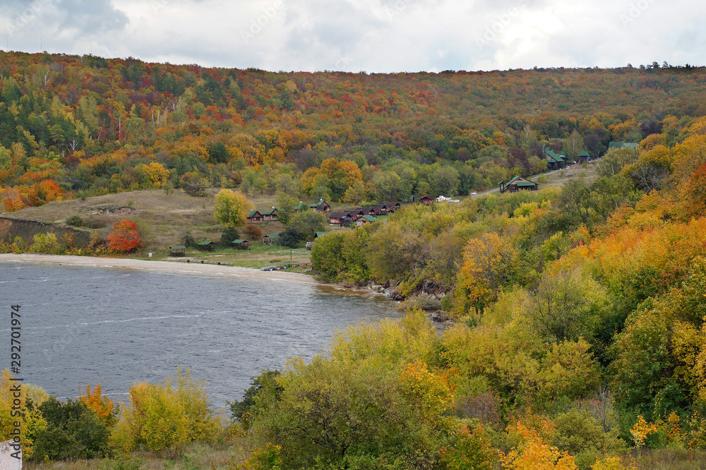 Autumn landscape: trees in different shades of gold, orange, green. On the shore of the lake there are tourist houses.