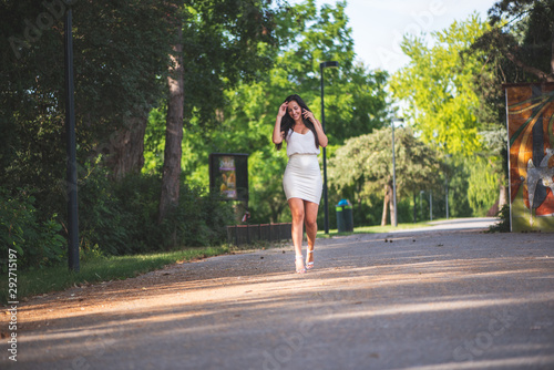 Businesswoman is walking through the park and talking on the phone. Woman in white top and skirt on the phone on a sunny day in the park with business customers. Smiling woman on the phone