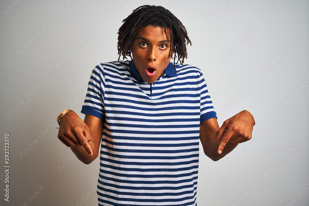 Afro man with dreadlocks wearing striped blue polo standing over isolated white background Pointing down with fingers showing advertisement, surprised face and open mouth Afro man with dreadlocks wearing striped blue polo standing over isolated white background Pointing down with fingers showing advertisement, surprised face and open mouth