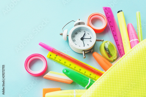 A white small alarm clock on a pencil case and pens, erasers, pencils, linear and multi-colored adhesive tape falling out of it...