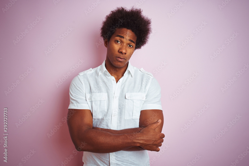 Young american man with afro hair wearing white shirt standing over isolated pink background skeptic and nervous, disapproving expression on face with crossed arms. Negative person. Young american man with afro hair wearing white shirt standing over isolated pink background skeptic and nervous, disapproving expression on face with crossed arms. Negative person.