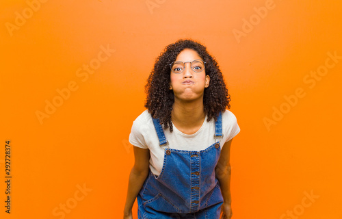 young pretty black woman with a goofy, crazy, surprised expression, puffing cheeks, feeling stuffed, fat and full of food against orange wall photo