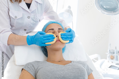 A woman  a professional doctor  a beautician cleanses the patient s face with sponges before applying the mask for skin care