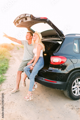 Young pretty couple pointing by hand smiling girlfriend sitting on car trunk in rural field © F8  \ Suport Ukraine