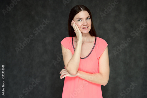 Portrait of a pretty young brunette woman with combed hair on a gray background in a pink blouse. It is in different poses. Looks straight at the camera. Slim figure.