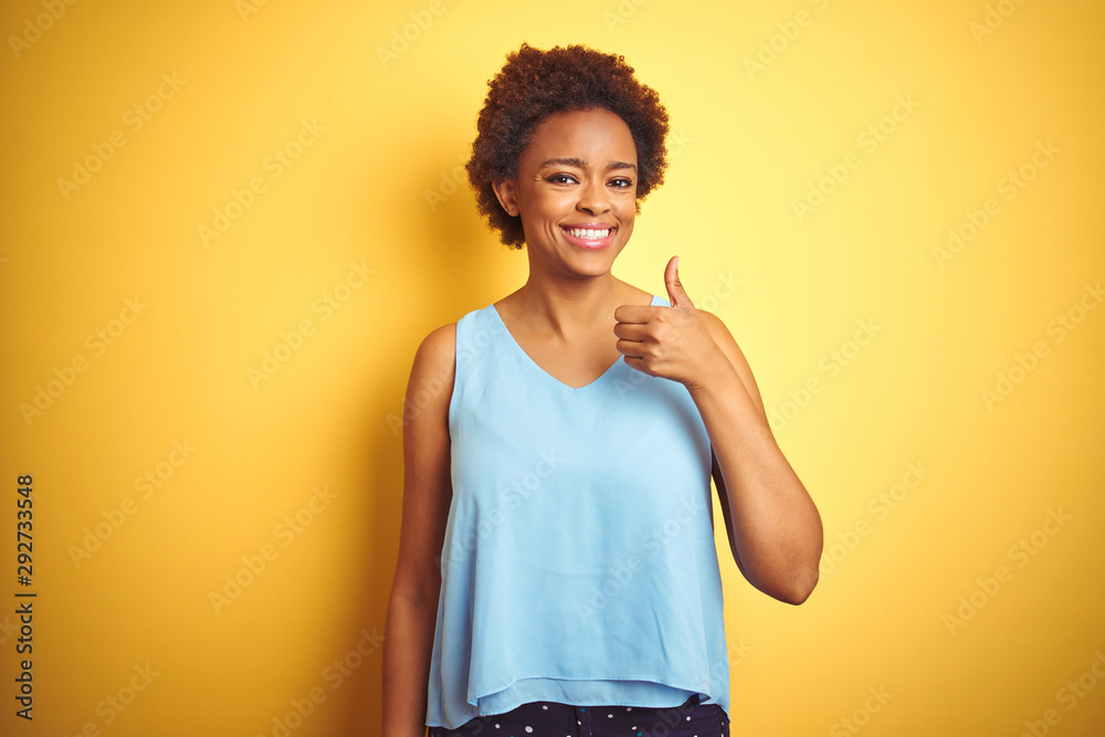 Beautiful african american woman wearing elegant shirt over isolated yellow background doing happy thumbs up gesture with hand. Approving expression looking at the camera showing success. Beautiful african american woman wearing elegant shirt over isolated yellow background doing happy thumbs up gesture with hand. Approving expression looking at the camera showing success.
